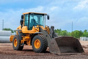 construction site with wheel loader in foreground, sewer pipe and tree line in background, sunny day, no people, machinery on dirt with bucket down on ground, blue sky with some clouds